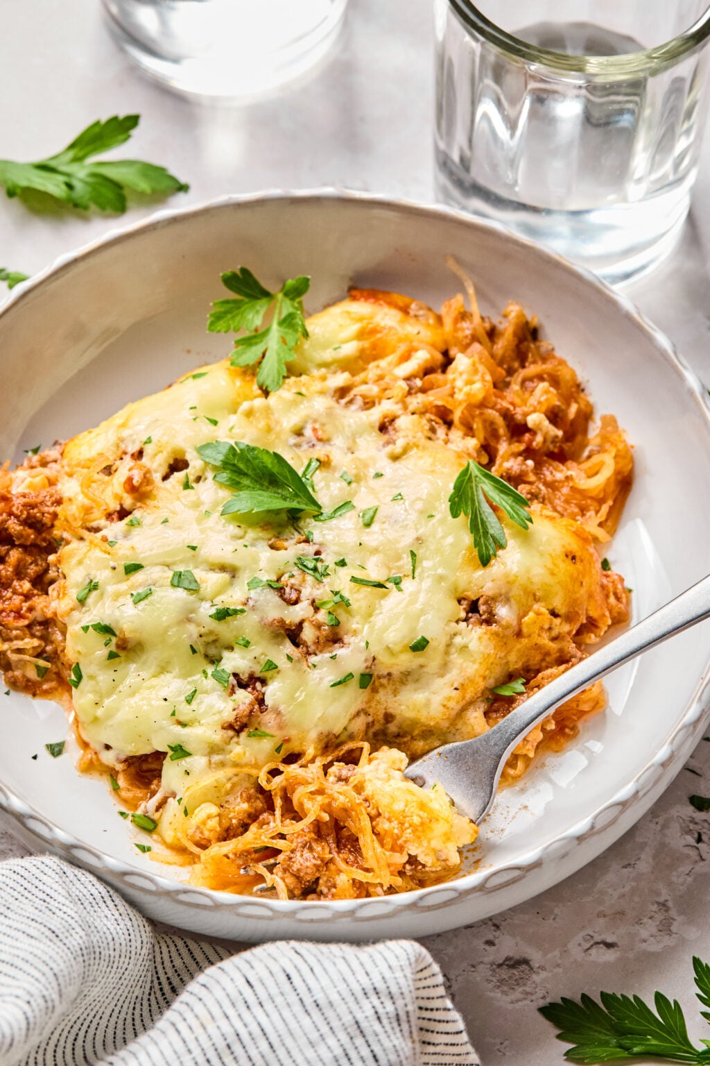 A plate of baked cheesy casserole with spaghetti squash, garnished with parsley, sits next to a fork, a striped napkin, and a glass of water.