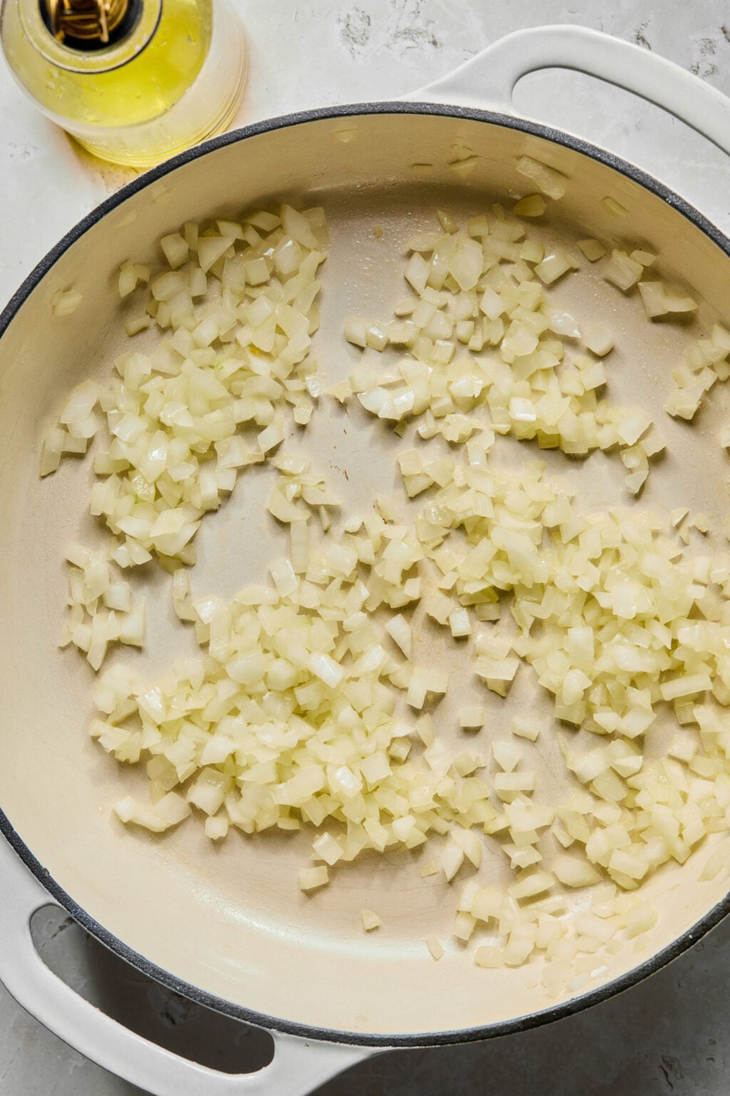 Chopped onions sautéing in a white enameled pan with a bottle of olive oil nearby.