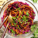 A glass bowl filled with shredded red cabbage, carrots, cilantro, and green onions, with salad tongs resting inside and various ingredients scattered nearby.