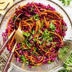 A glass bowl filled with shredded red cabbage, carrots, cilantro, and green onions, with salad tongs resting inside and various ingredients scattered nearby.