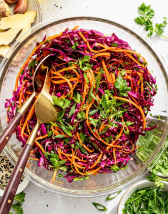 A glass bowl filled with shredded red cabbage, carrots, cilantro, and green onions, with salad tongs resting inside and various ingredients scattered nearby.