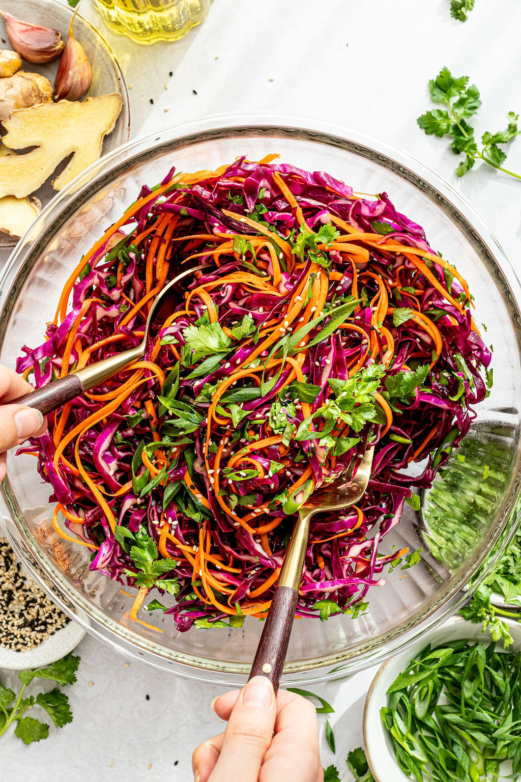 A person tosses a colorful salad with shredded red cabbage, carrots, cilantro, and sesame seeds in a clear glass bowl.