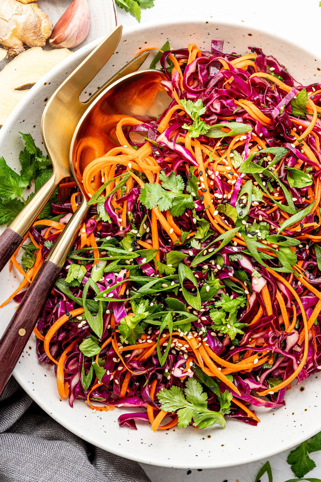 A bowl of colorful Asian slaw with shredded red cabbage, carrots, cilantro, green onions, and sesame seeds, served with wooden salad utensils.