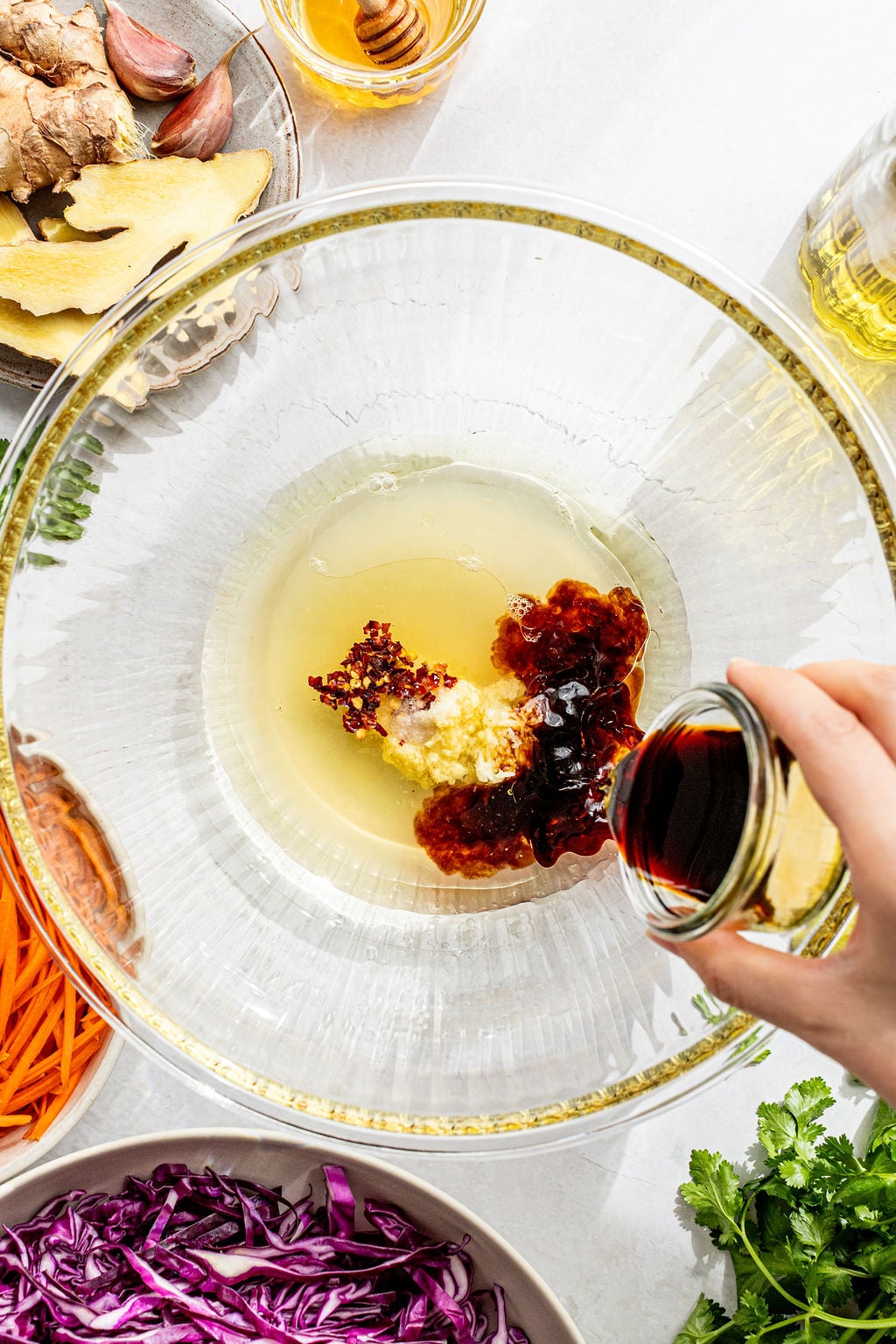 A hand pours soy sauce into a bowl containing minced garlic, chili flakes, and other sauces, surrounded by vegetables and seasoning ingredients.