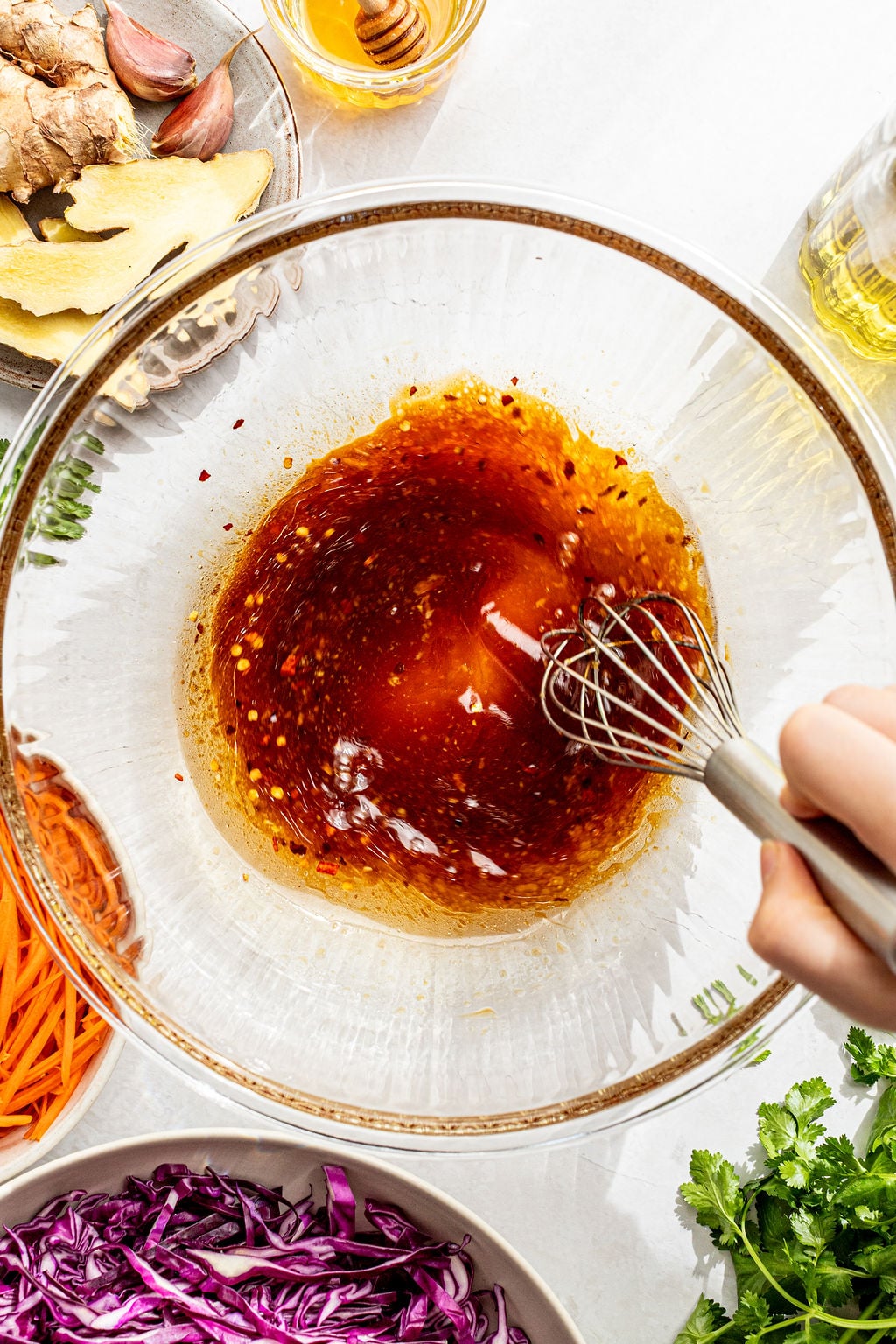 A hand uses a whisk to mix a reddish-brown sauce in a glass bowl, surrounded by ingredients like shredded carrots, purple cabbage, ginger, and cilantro.
