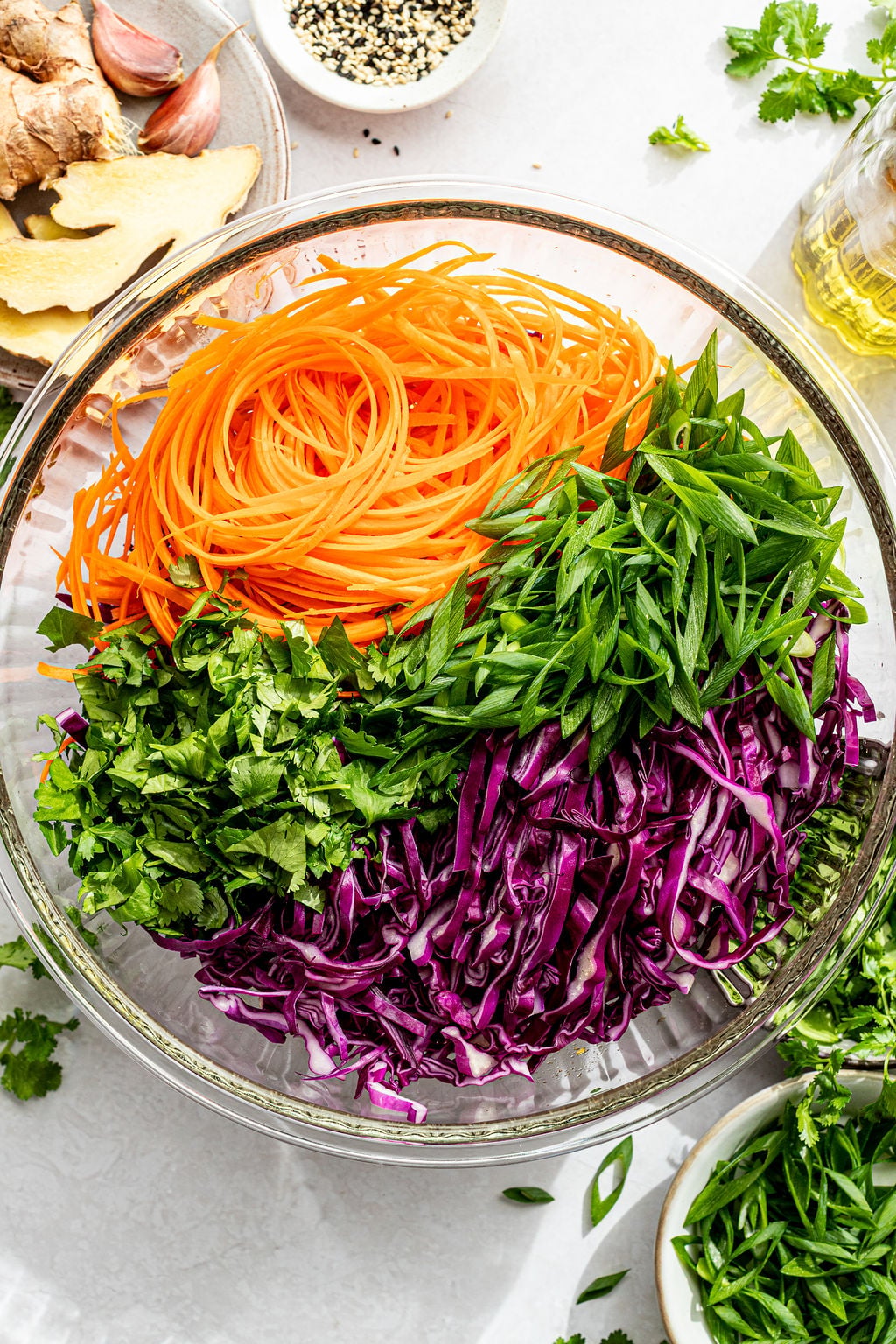 A glass bowl filled with shredded carrots, sliced green onions, chopped cilantro, and shredded purple cabbage, arranged in separate sections.