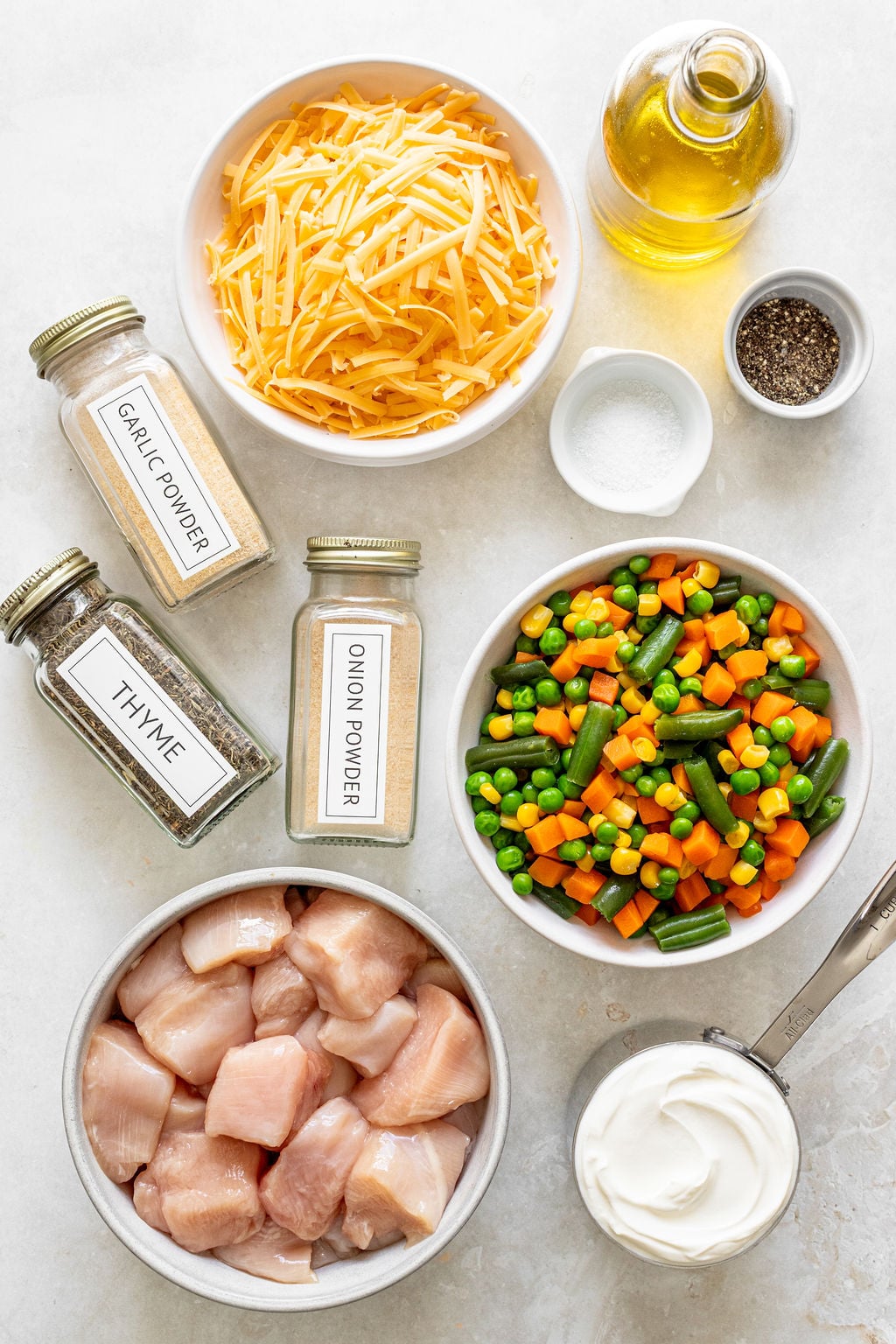 Top-down view of ingredients including cubed raw chicken, shredded cheese, mixed frozen vegetables, seasonings, olive oil, salt, pepper, and sour cream arranged in bowls and jars.