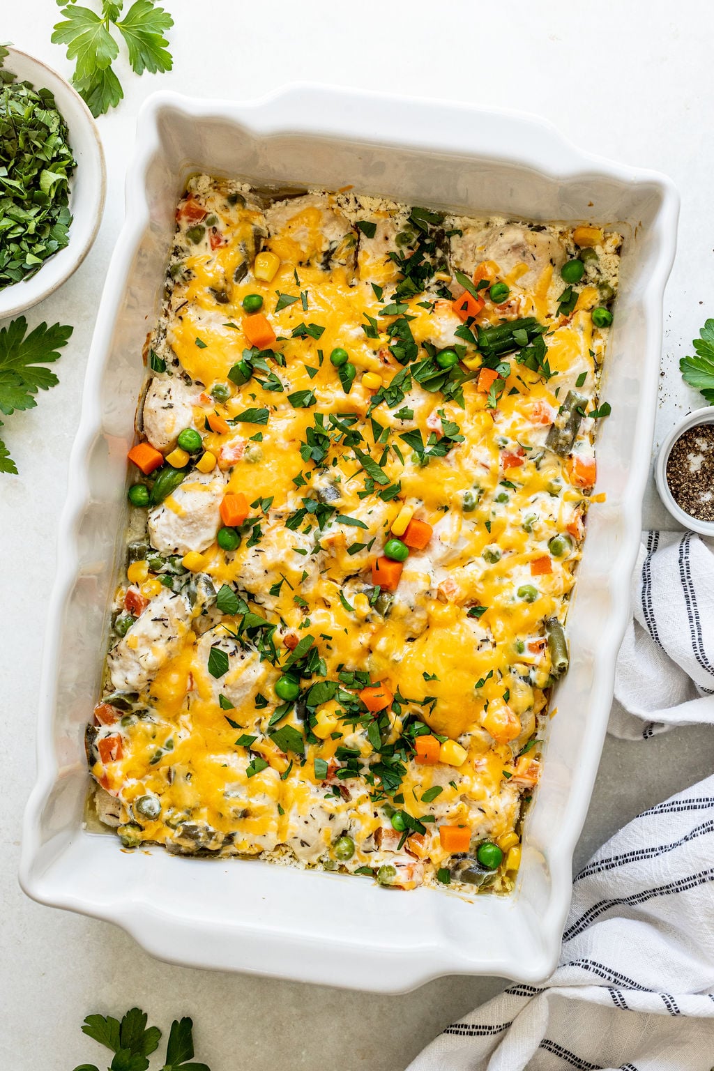 A white casserole dish filled with a baked mixture of diced chicken, mixed vegetables, and melted cheese, garnished with chopped herbs.