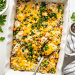 A baked casserole in a white dish topped with melted cheese, mixed vegetables, and herbs, with a serving spoon and a side bowl of chopped parsley.