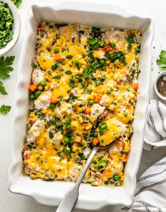 A baked casserole in a white dish topped with melted cheese, mixed vegetables, and herbs, with a serving spoon and a side bowl of chopped parsley.