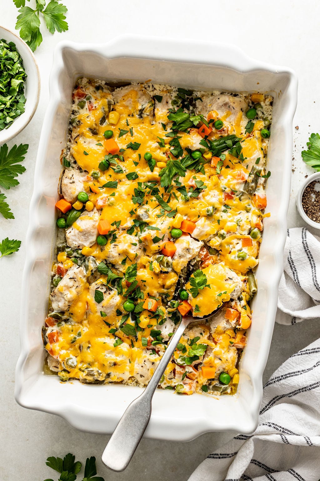 A baked casserole in a white dish topped with melted cheese, mixed vegetables, and herbs, with a serving spoon and a side bowl of chopped parsley.