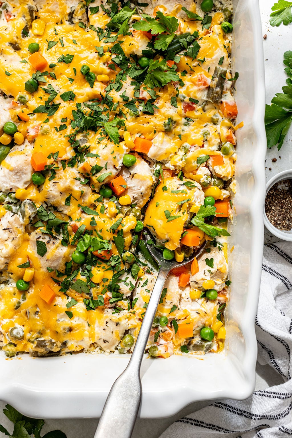 A baked casserole with melted cheese, diced chicken, peas, carrots, corn, and fresh herbs, in a white dish with a serving spoon.