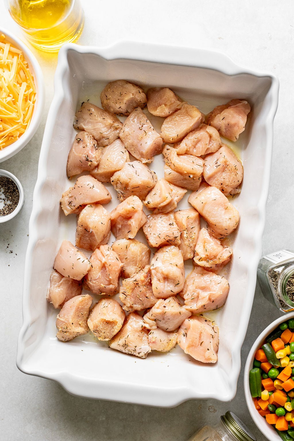 Raw, seasoned chicken pieces arranged in a white baking dish, surrounded by bowls of shredded cheese, mixed vegetables, oil, and seasonings on a light surface.