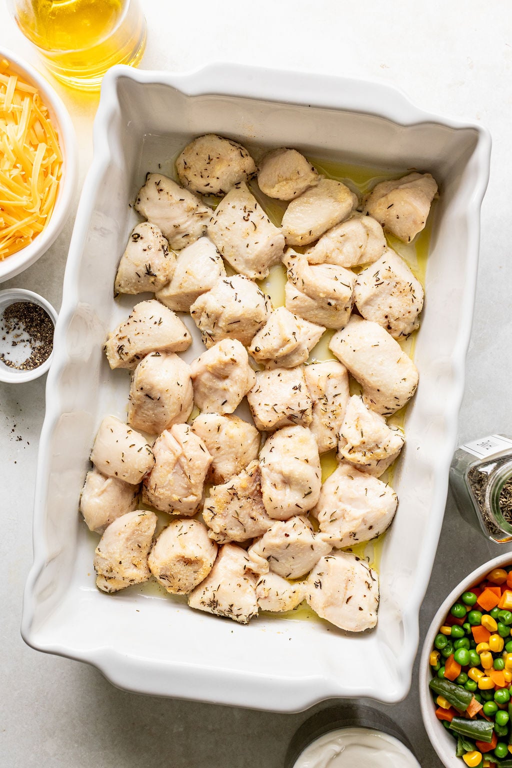 Raw chicken pieces seasoned with herbs arranged in a white baking dish, surrounded by bowls of shredded cheese, mixed vegetables, black pepper, and oil.