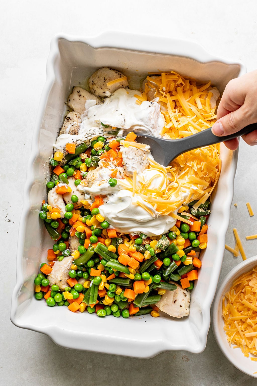 A hand stirs shredded cheese, sour cream, and mixed vegetables with chicken pieces in a white baking dish.