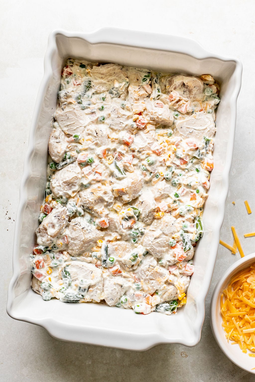 A rectangular baking dish filled with an uncooked creamy mixture of chopped vegetables, shredded cheese, and chicken. A small bowl of shredded cheese sits nearby.
