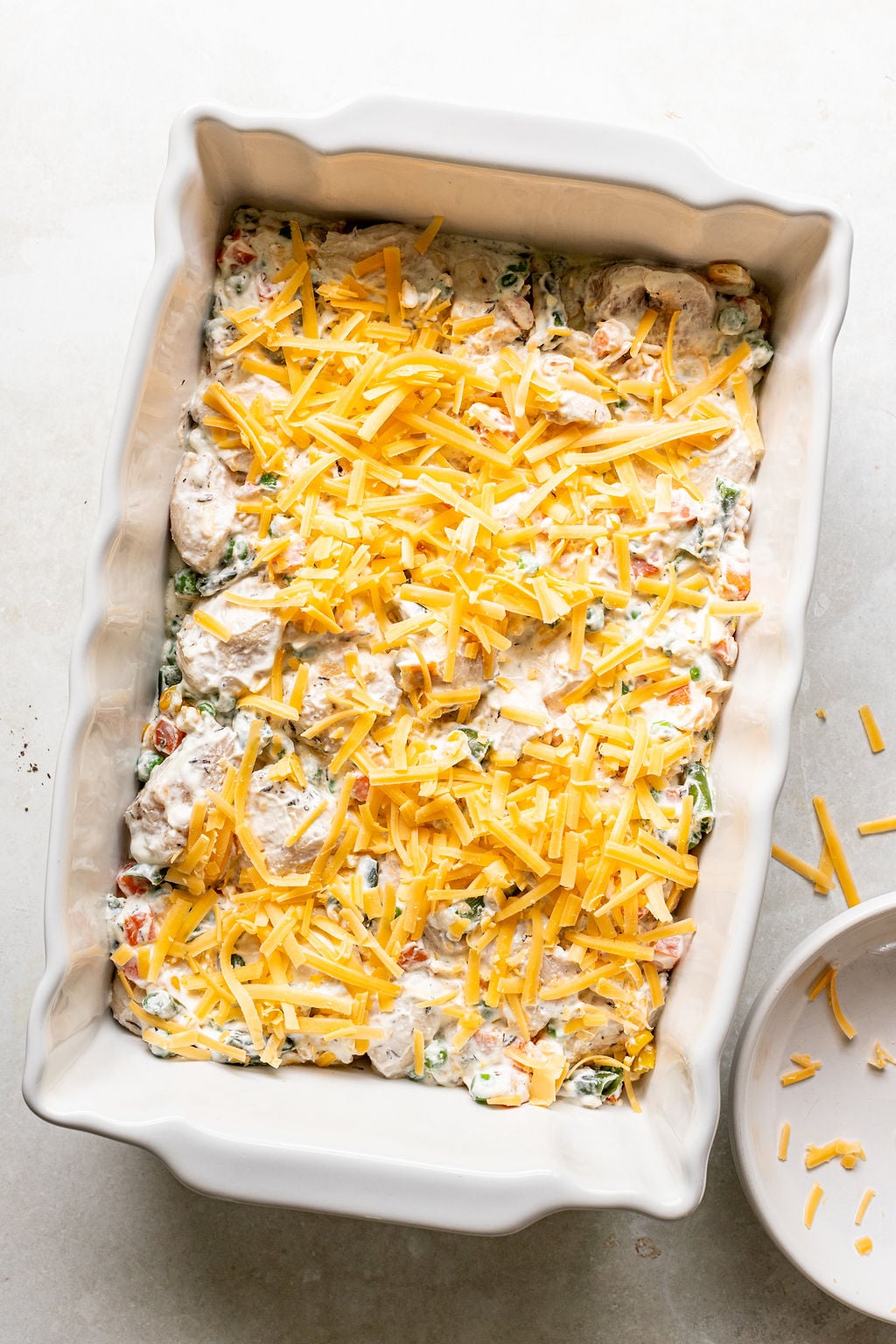 Rectangular baking dish filled with a creamy mixture of vegetables and shredded cheddar cheese, ready to be baked. A small bowl with extra cheese is beside the dish.