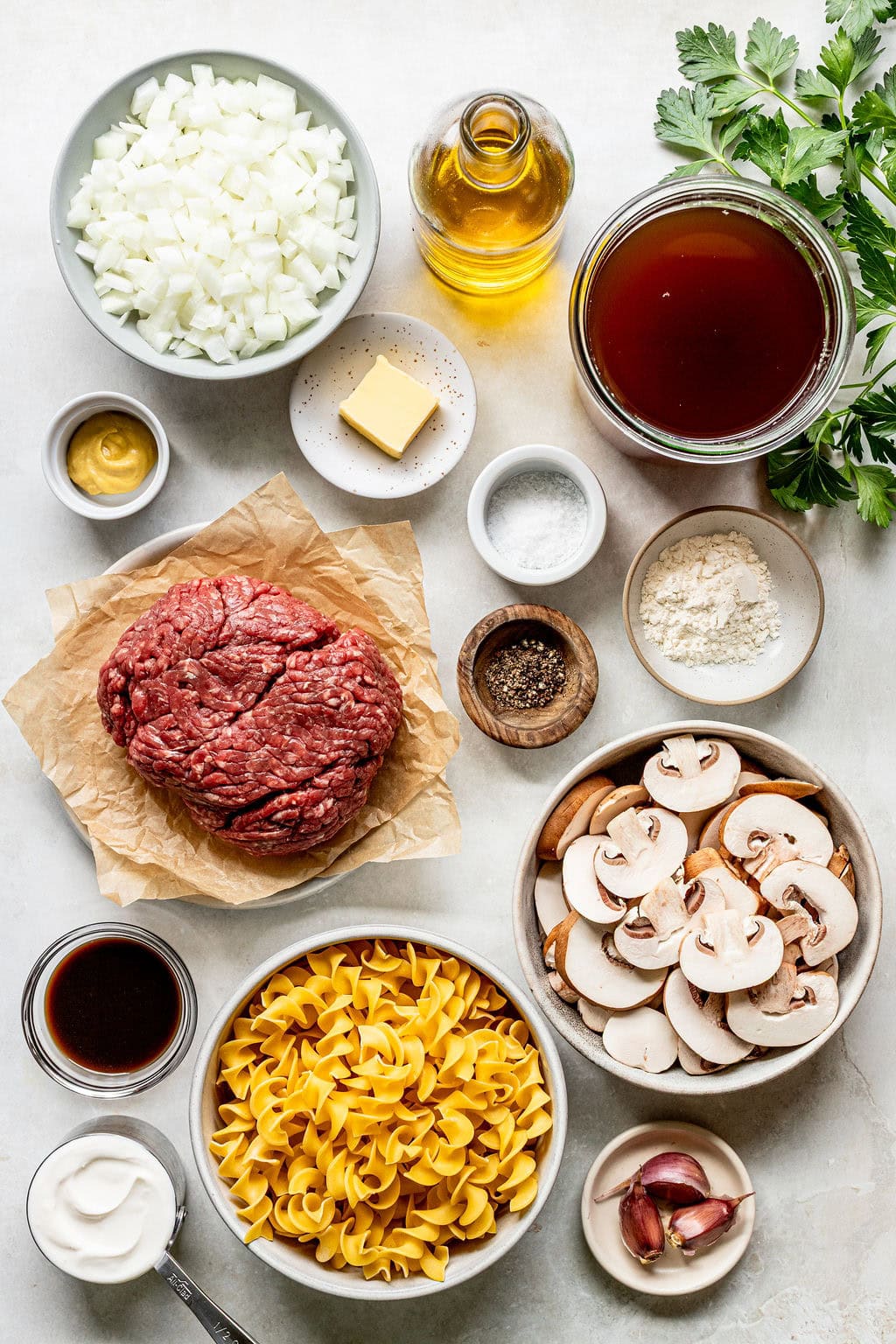 Top view of ingredients for beef stroganoff, including ground beef, egg noodles, mushrooms, onion, garlic, broth, sour cream, mustard, butter, flour, oil, parsley, salt, and pepper.