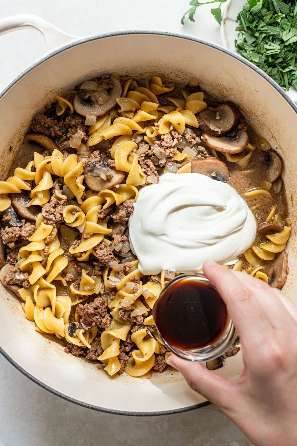 A hand pours a small glass of dark liquid into a pot containing cooked egg noodles, ground meat, mushrooms, onions, and a large dollop of sour cream. Fresh herbs are nearby.