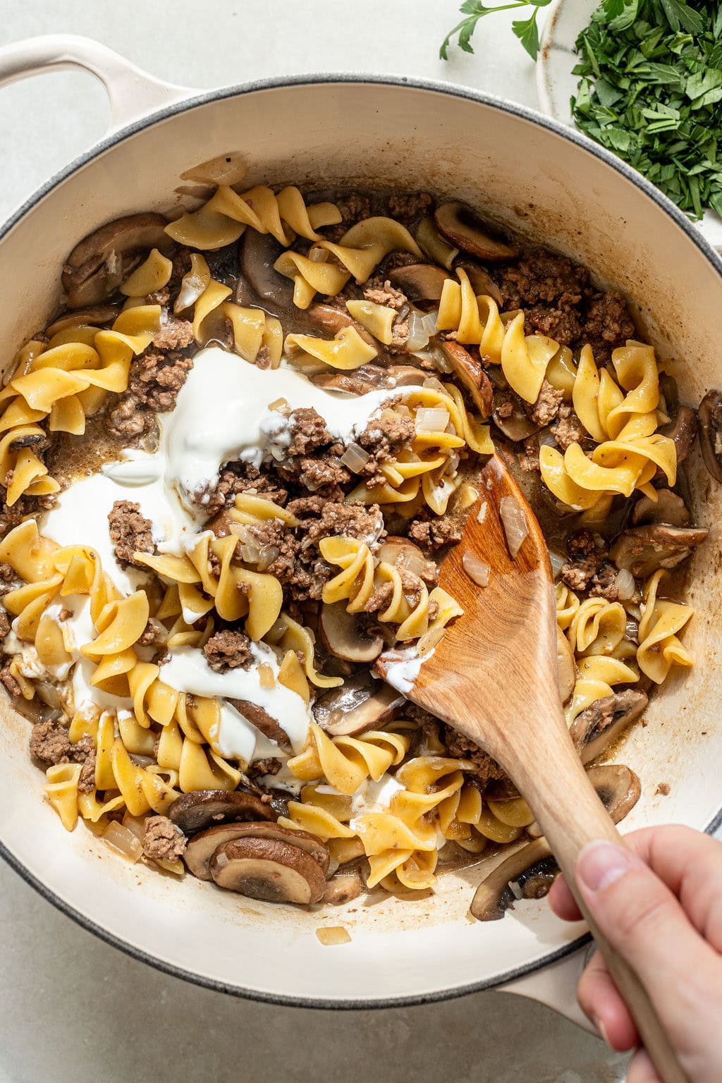 A hand stirs cooked noodles, ground beef, mushrooms, and a dollop of sour cream in a large white pot. Chopped herbs are on the side.