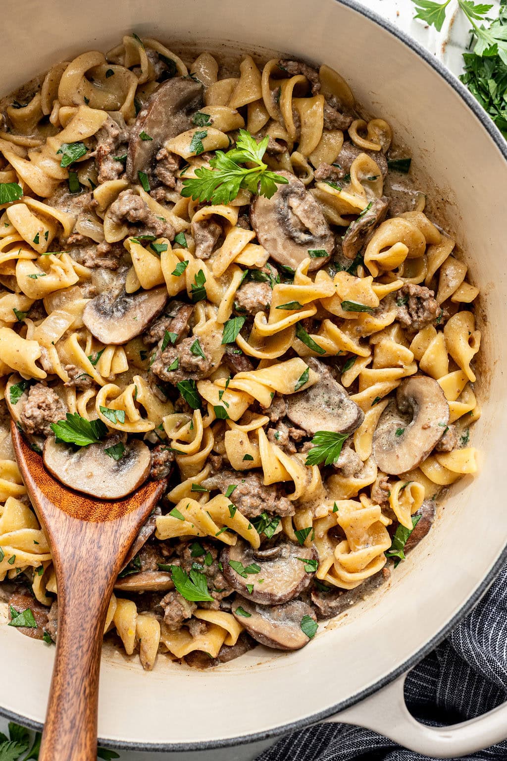 A pot of beef stroganoff with egg noodles, mushrooms, and parsley, stirred with a wooden spoon.