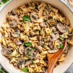 A large pan of cooked egg noodles with sliced mushrooms, ground meat, and a creamy sauce, garnished with chopped parsley and served with a wooden spoon.