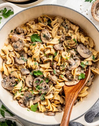 A large pan of cooked egg noodles with sliced mushrooms, ground meat, and a creamy sauce, garnished with chopped parsley and served with a wooden spoon.