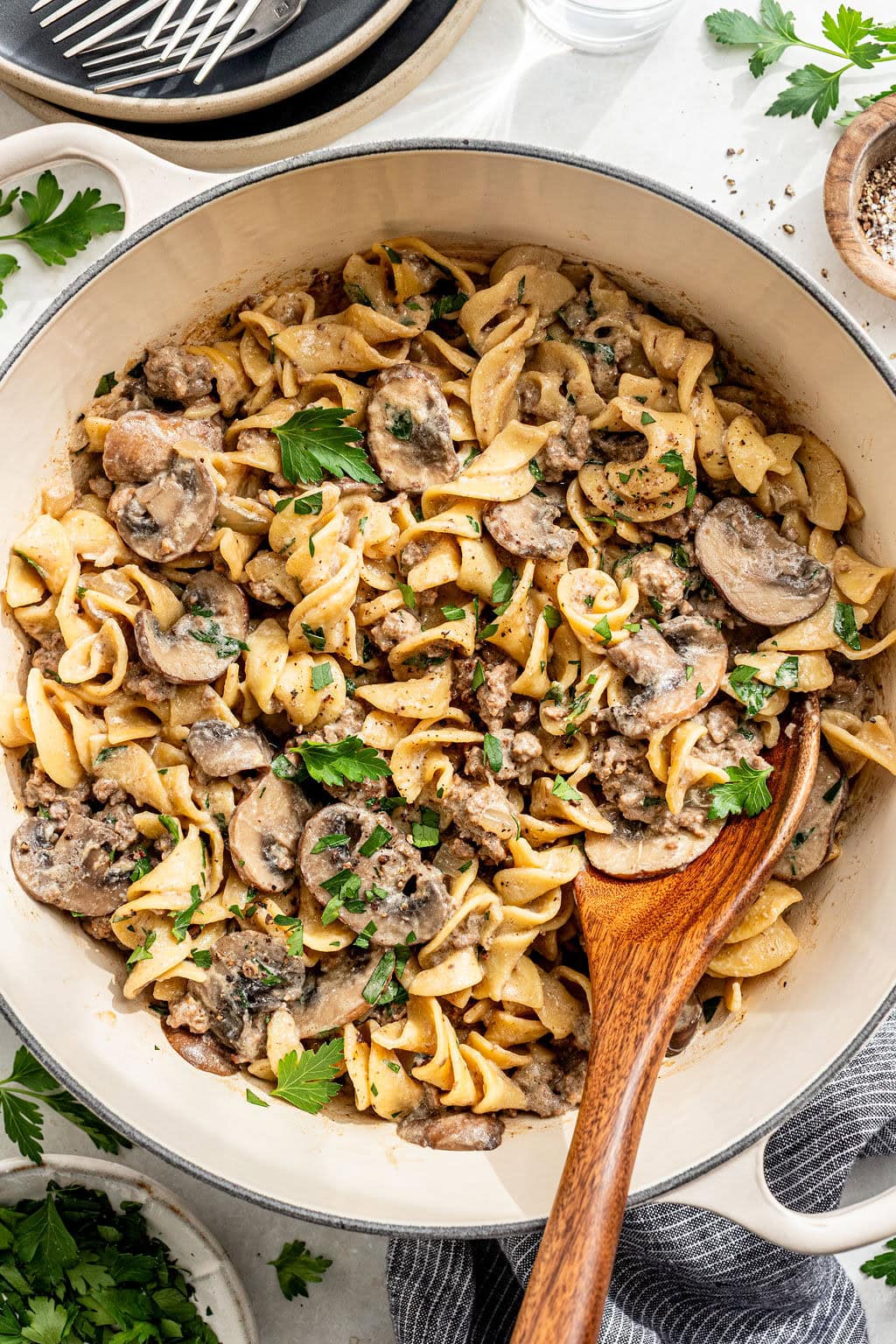 A large pan of cooked egg noodles with sliced mushrooms, ground meat, and a creamy sauce, garnished with chopped parsley and served with a wooden spoon.