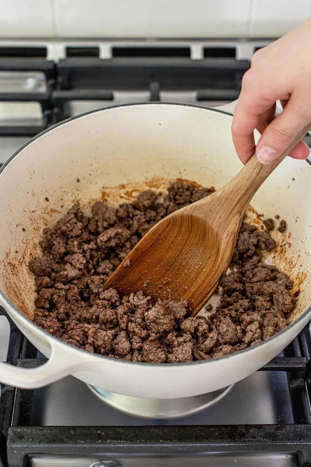 A hand stirs cooked ground beef in a white pot with a wooden spoon on a stovetop.