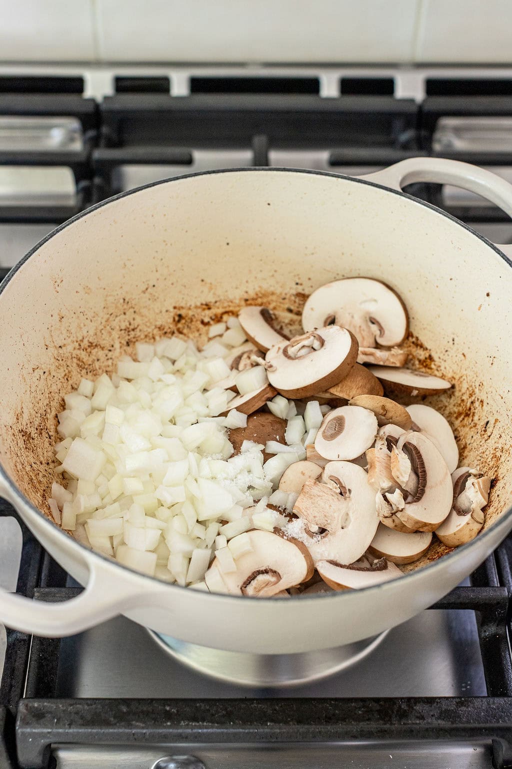 Chopped onions and sliced mushrooms in a white pot on a stove, beginning to sauté.