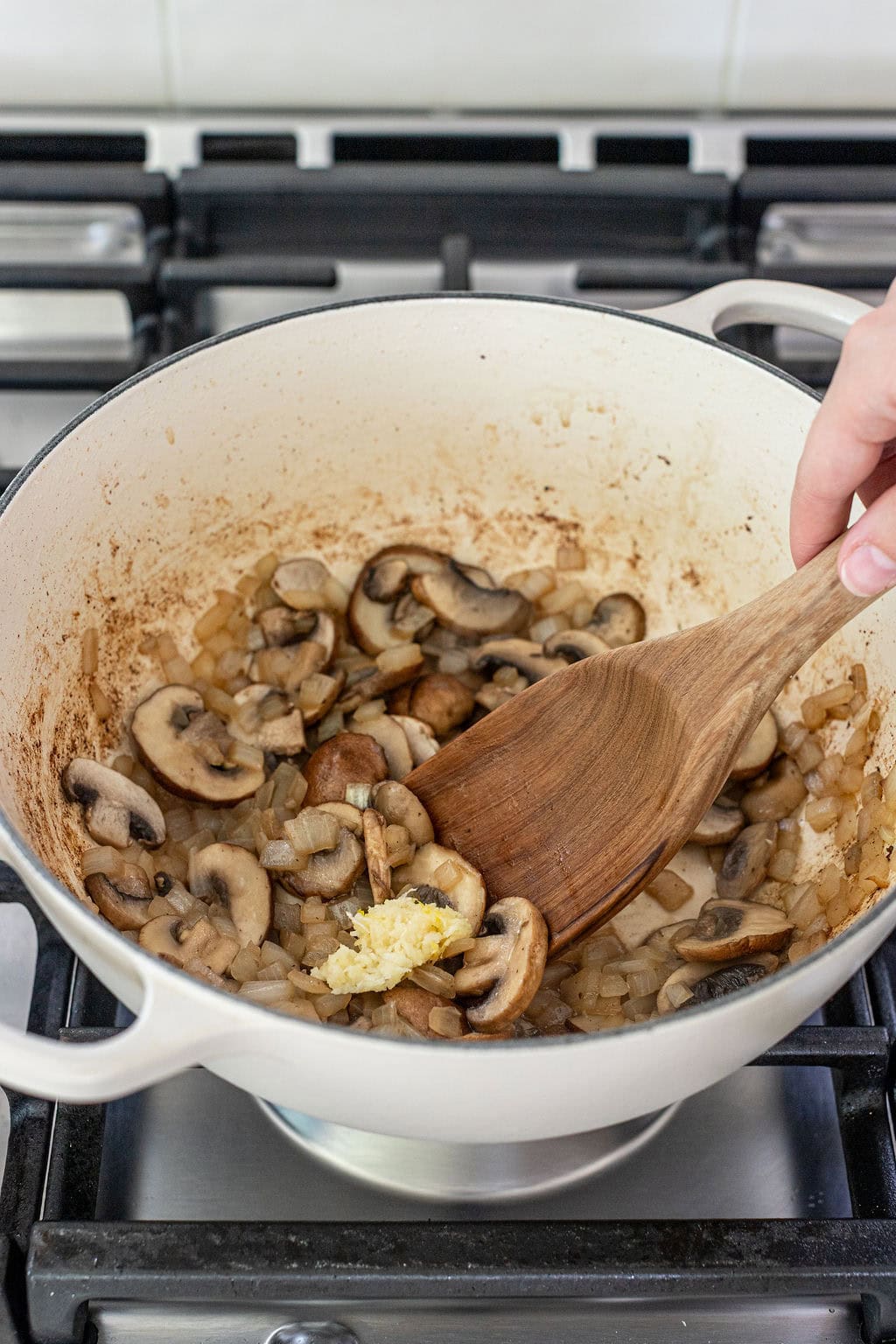 A hand stirs chopped onions, mushrooms, and minced garlic in a white pot on a stovetop.