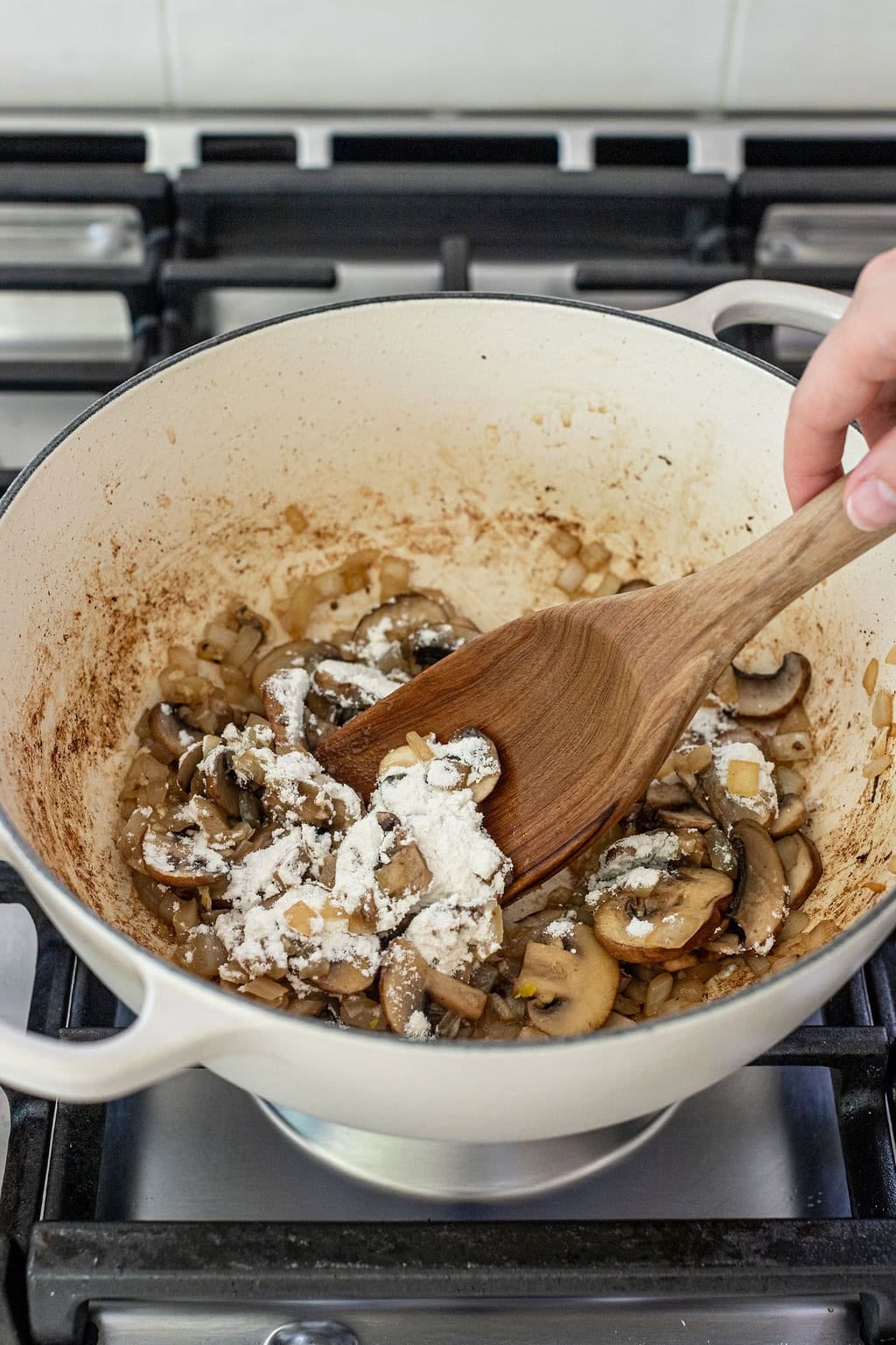A hand stirs chopped onions, mushrooms, and flour in a white pot on a gas stove.