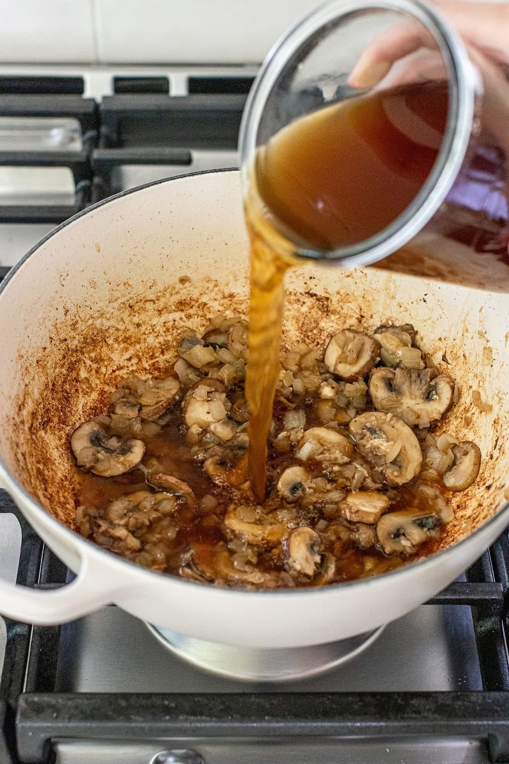 A person pours brown broth into a white pot containing sliced mushrooms and onions on a stovetop.