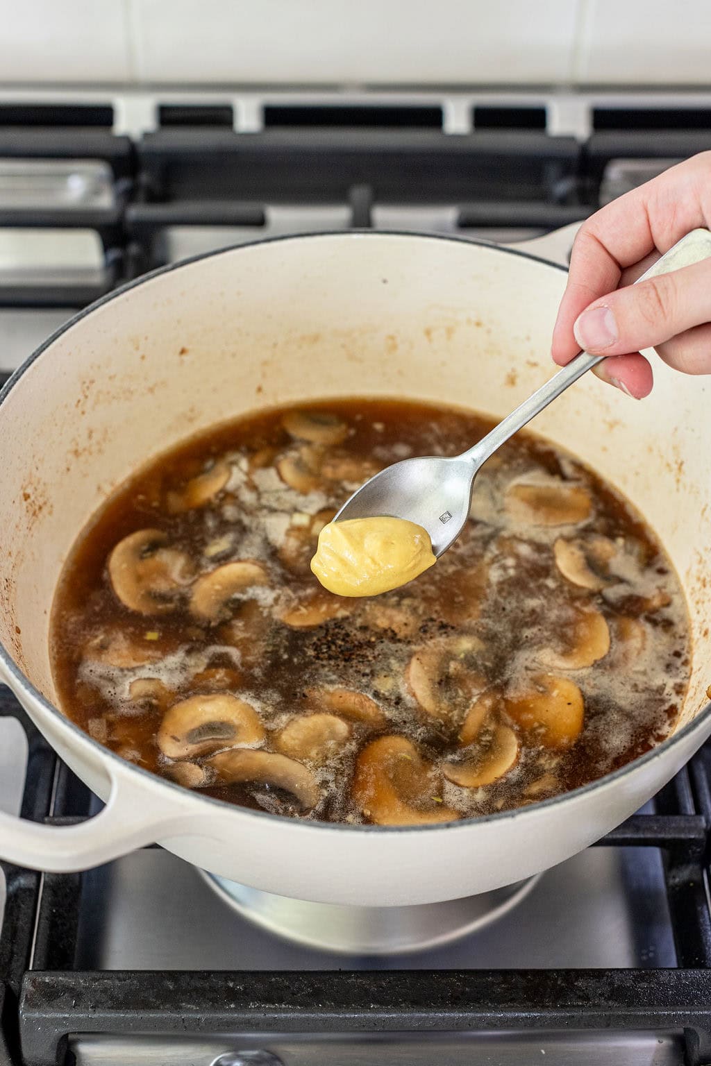 A hand holds a spoonful of mustard over a pot of simmering broth with sliced mushrooms on a stove.