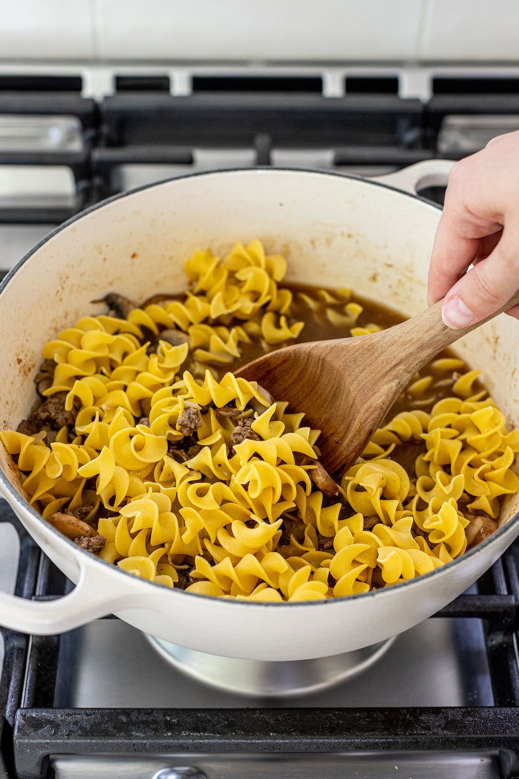 A hand stirs uncooked rotini pasta and beef in a white pot on a stove.