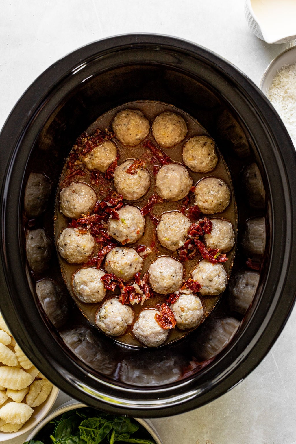 Slow cooker filled with cooked meatballs in sauce, topped with chopped sun-dried tomatoes. Ingredients like gnocchi, spinach, and shredded cheese are placed nearby.