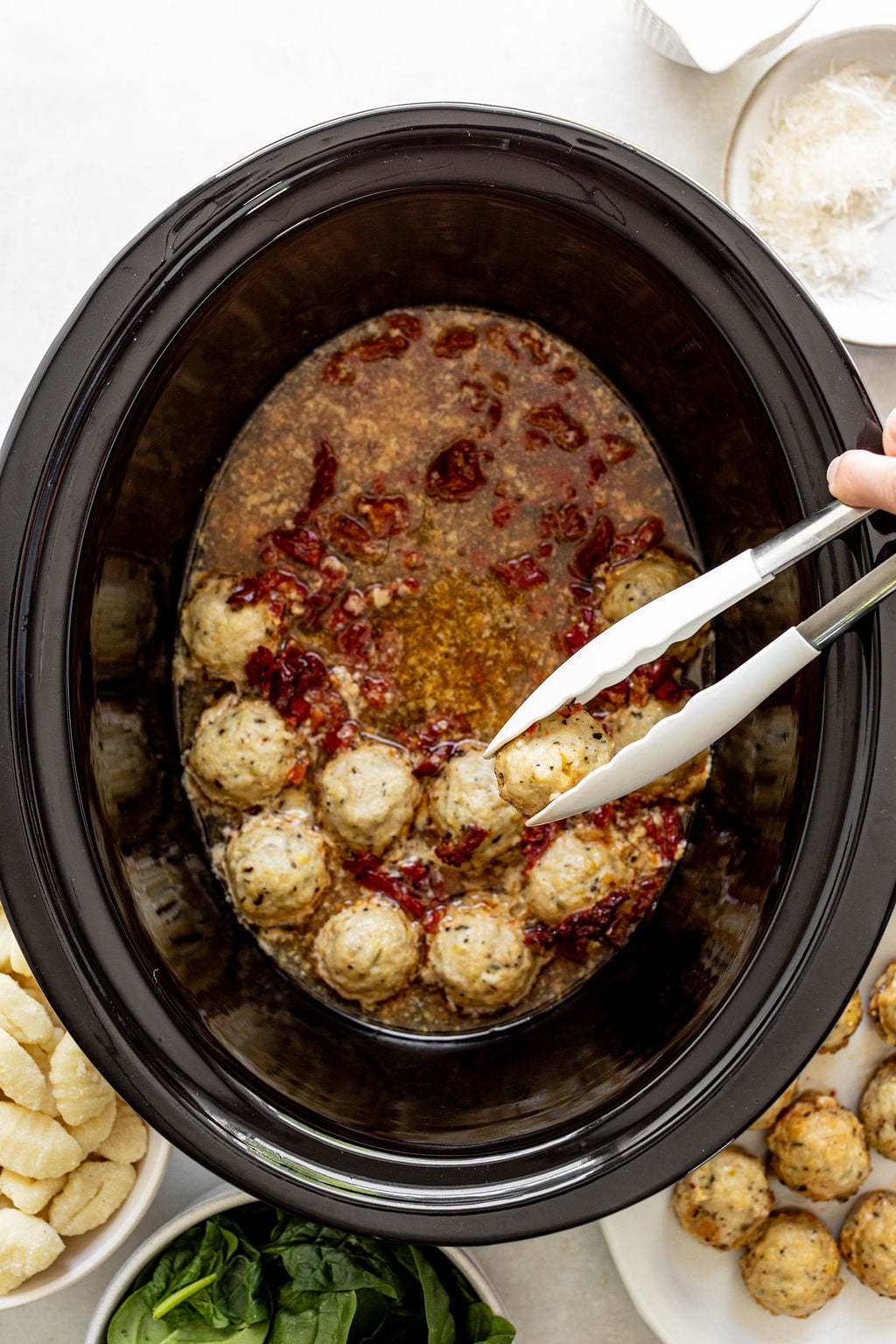 A slow cooker filled with cooked chicken meatballs, sauce, and sun-dried tomatoes with tongs lifting a meatball; gnocchi, spinach, and Parmesan on the side.