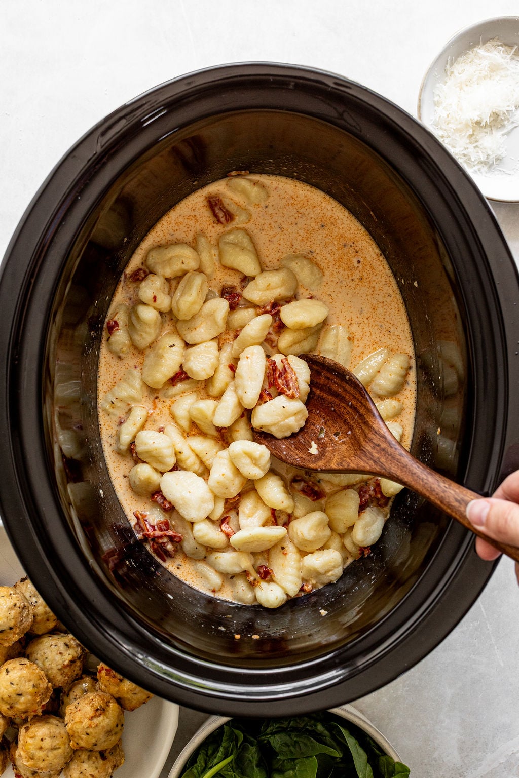 A hand stirs gnocchi, sun-dried tomatoes, and creamy sauce in a slow cooker with a wooden spoon. Bowls of spinach, meatballs, and grated cheese are nearby.