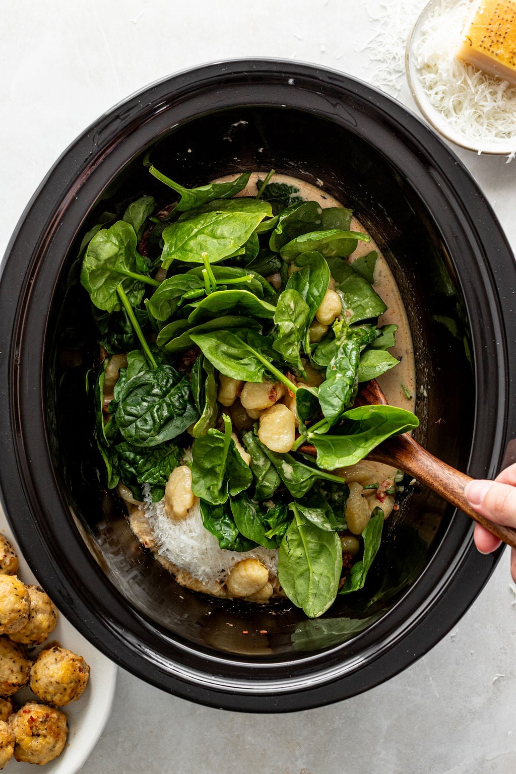 A hand stirs fresh spinach and gnocchi in a slow cooker with a wooden spoon. A bowl of grated cheese and a plate of cooked food are nearby.