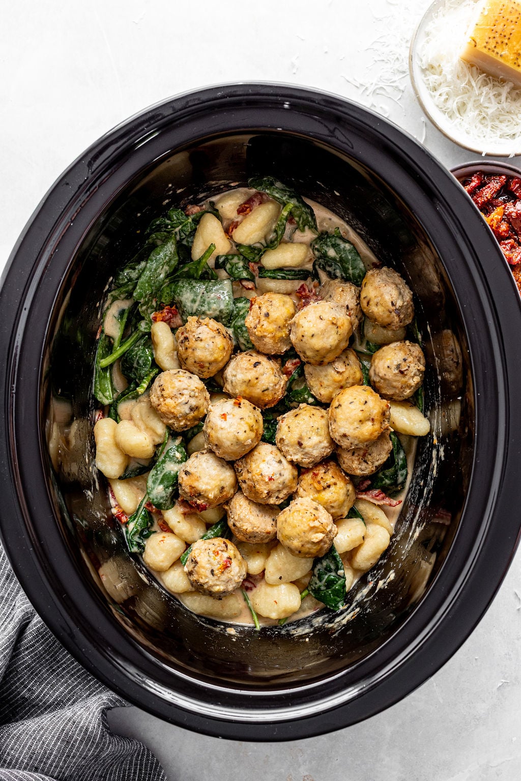 A slow cooker filled with gnocchi, spinach, sun-dried tomatoes, and meatballs in a creamy sauce. A bowl of grated cheese and sun-dried tomatoes are nearby.