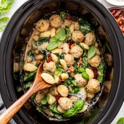 A slow cooker filled with creamy gnocchi, chicken meatballs, spinach, sun-dried tomatoes, and fresh basil, with a wooden spoon resting inside.