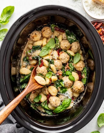 A slow cooker filled with creamy gnocchi, chicken meatballs, spinach, sun-dried tomatoes, and fresh basil, with a wooden spoon resting inside.