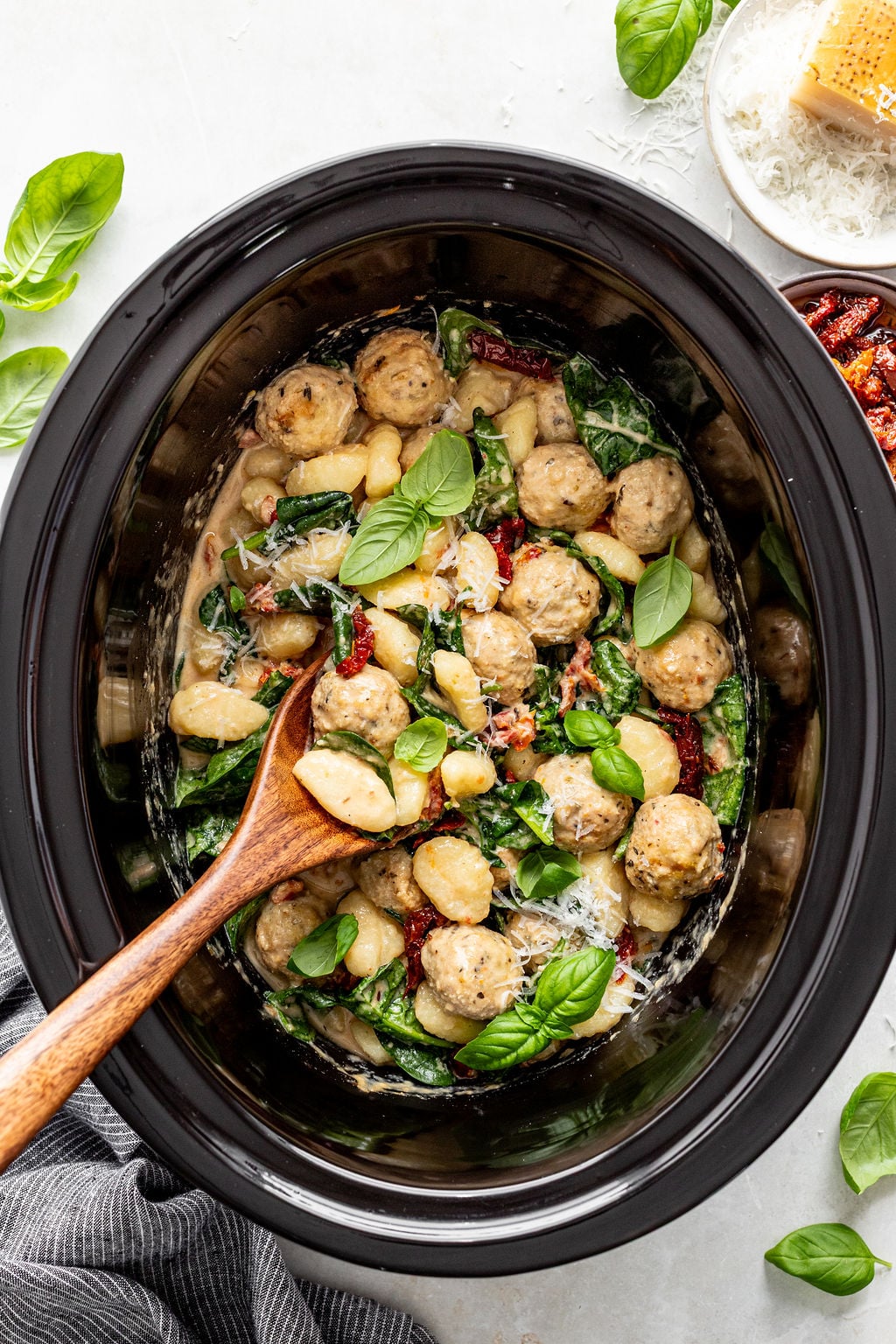 A slow cooker filled with creamy gnocchi, chicken meatballs, spinach, sun-dried tomatoes, and fresh basil, with a wooden spoon resting inside.