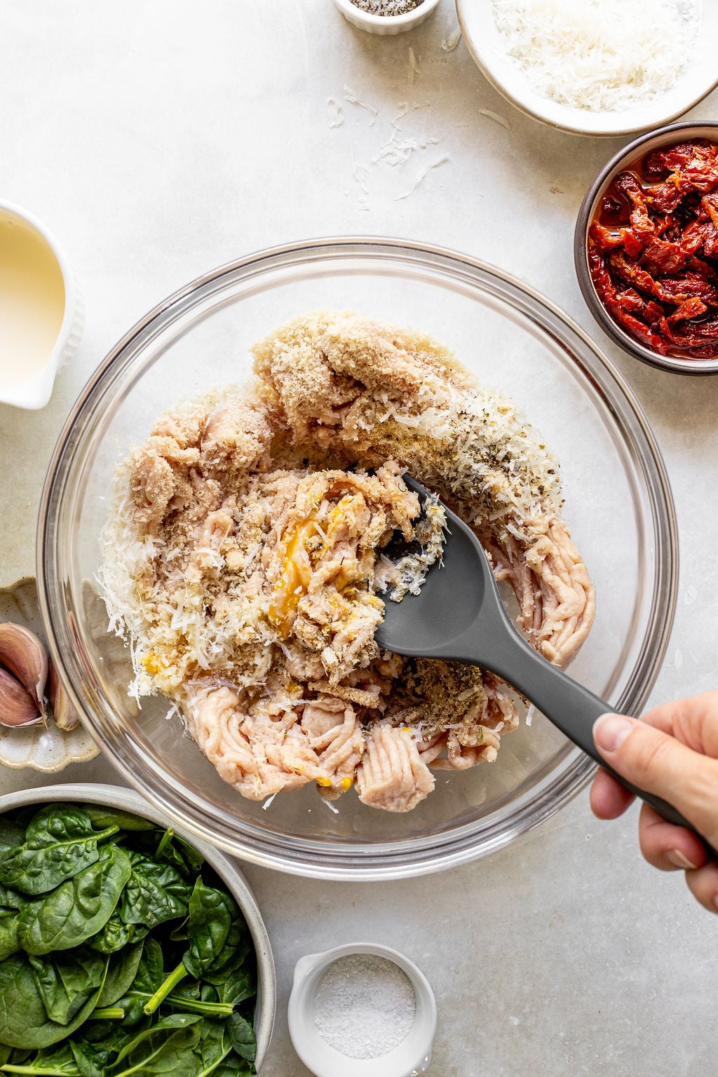 A hand mixes ground chicken, breadcrumbs, and cheese in a glass bowl with a black spatula; spinach, salt, sun-dried tomatoes, and cream are nearby.