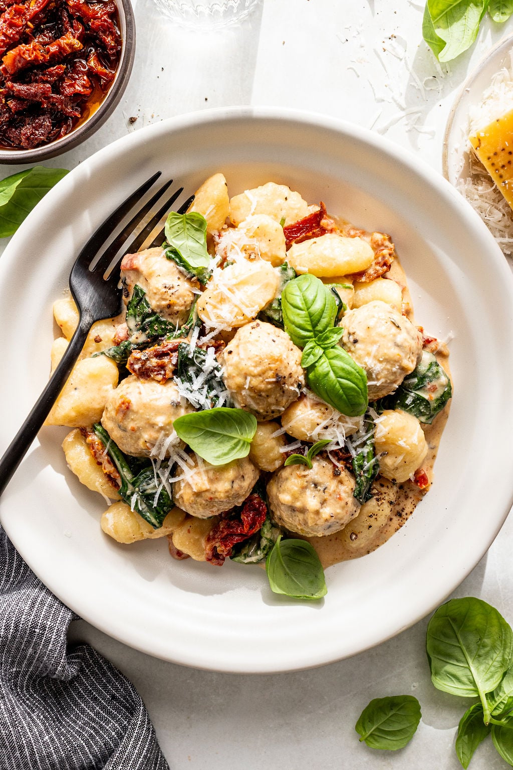 A plate of gnocchi with meatballs, sun-dried tomatoes, spinach, cream sauce, and grated cheese, garnished with fresh basil leaves. A black fork rests on the plate.