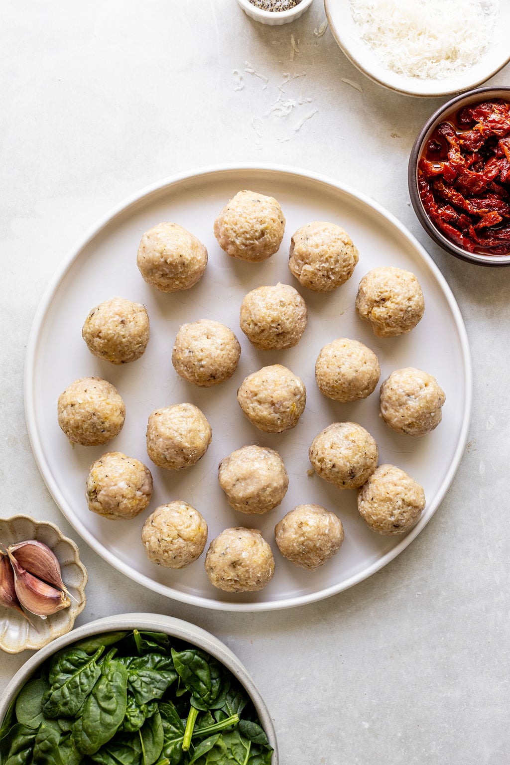 A white plate with 18 uncooked meatballs is surrounded by bowls of sun-dried tomatoes, shredded cheese, spinach, and garlic cloves on a light countertop.