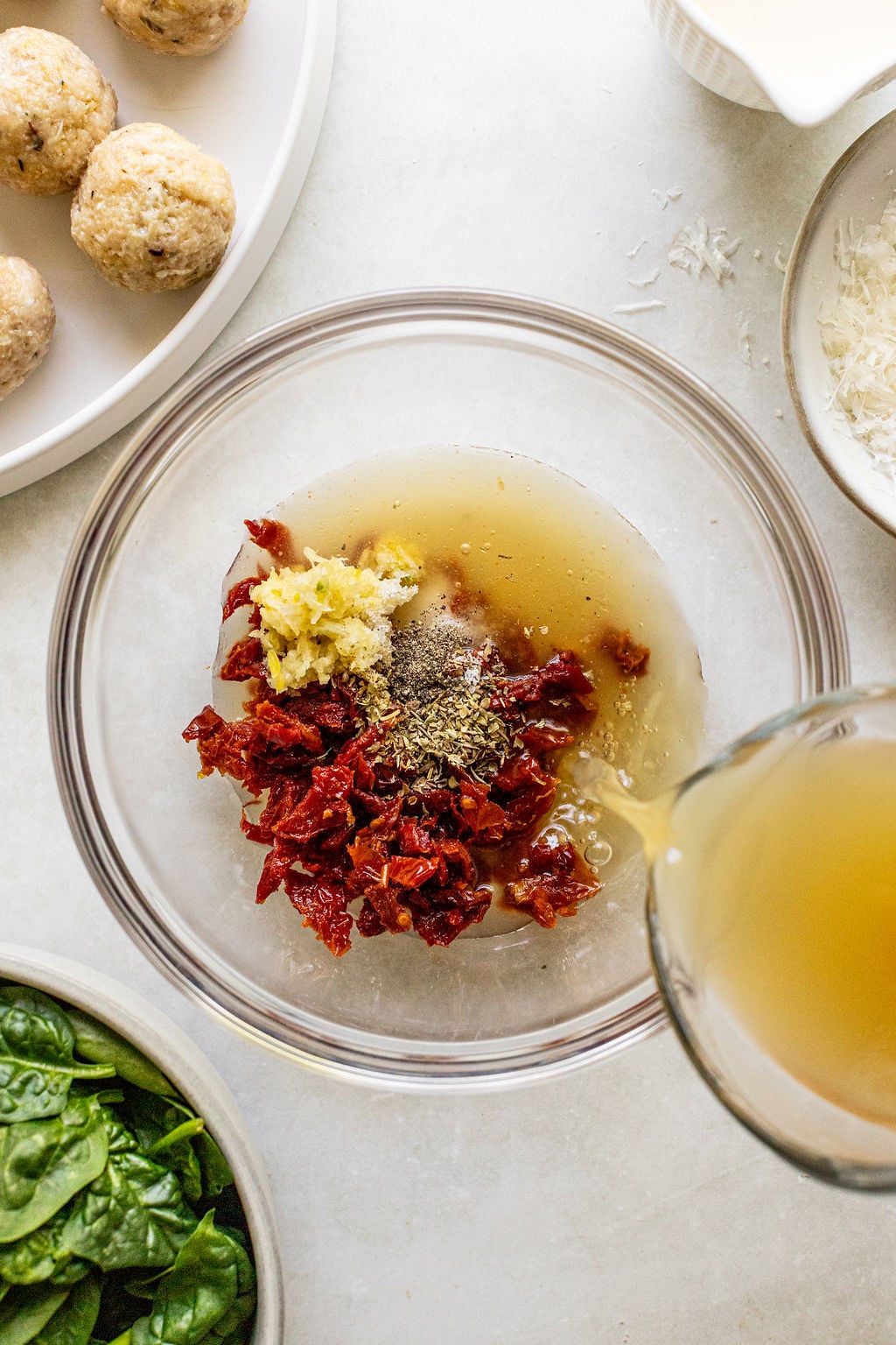 A glass bowl with sun-dried tomatoes, minced garlic, herbs, and liquid being poured in. Surrounding bowls contain spinach, meatballs, and grated cheese.