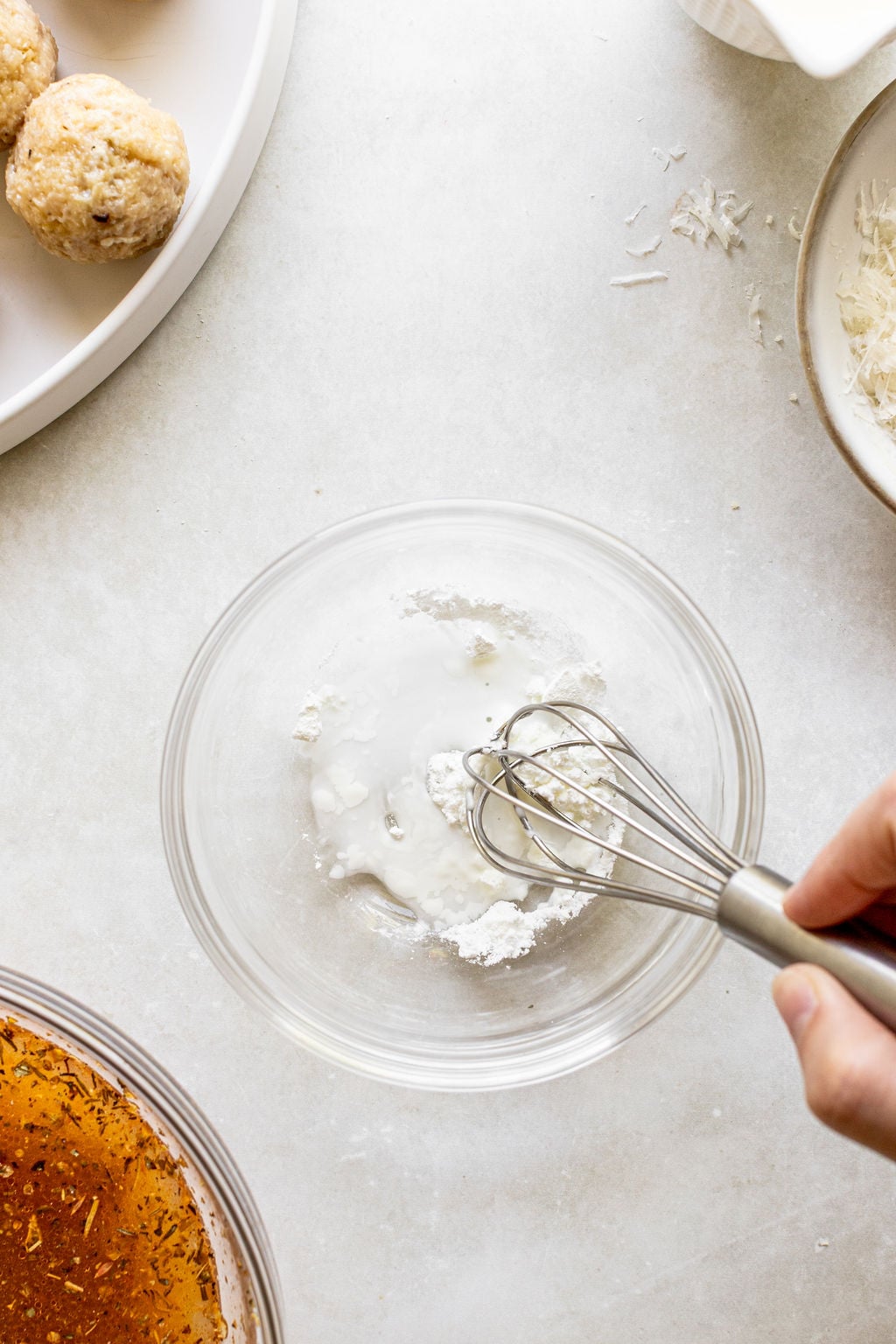 A hand whisking a white powder and liquid mixture in a glass bowl on a light countertop, with food ingredients nearby.