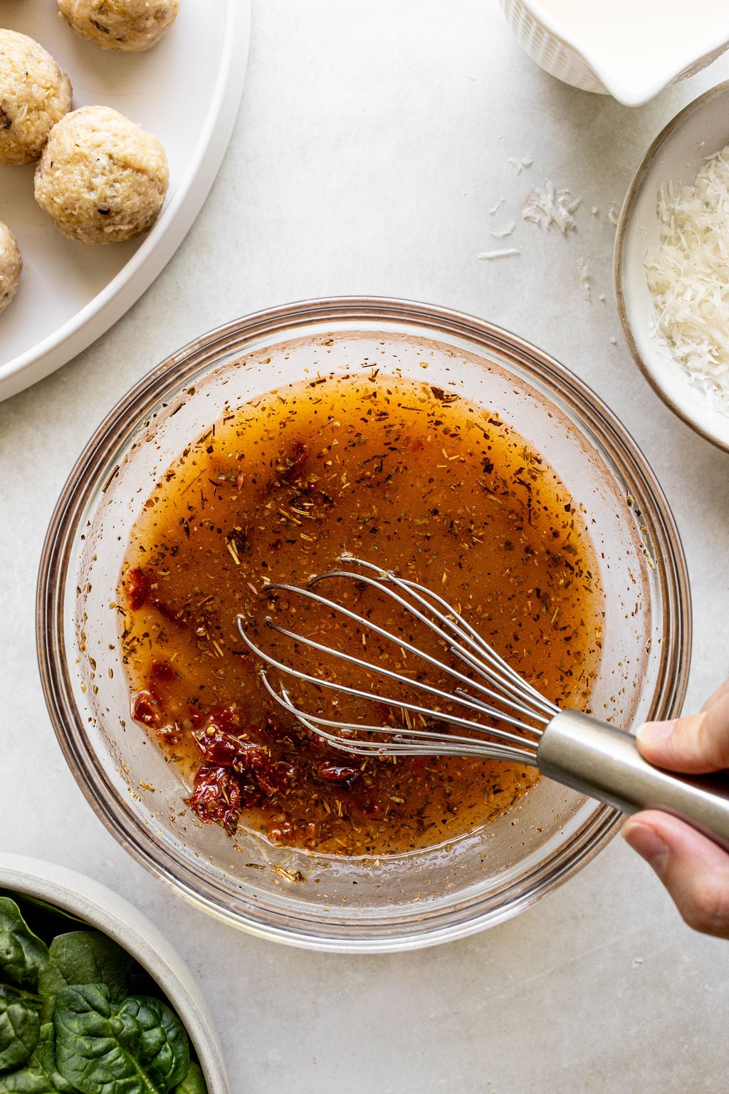 A hand whisks a reddish-brown seasoned liquid in a glass bowl, surrounded by plates of raw meatballs, shredded cheese, and fresh spinach.