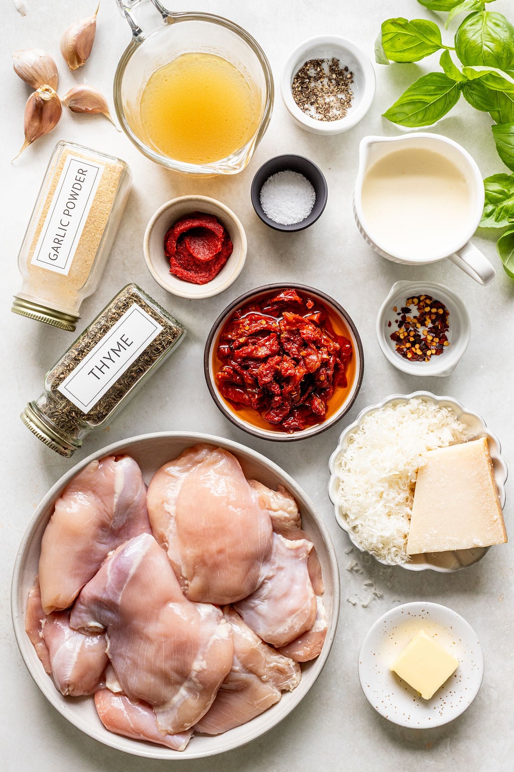 A flat lay of ingredients including raw chicken thighs, sun-dried tomatoes, grated cheese, cream, broth, butter, garlic, spices, salt, pepper, and fresh basil on a white surface.